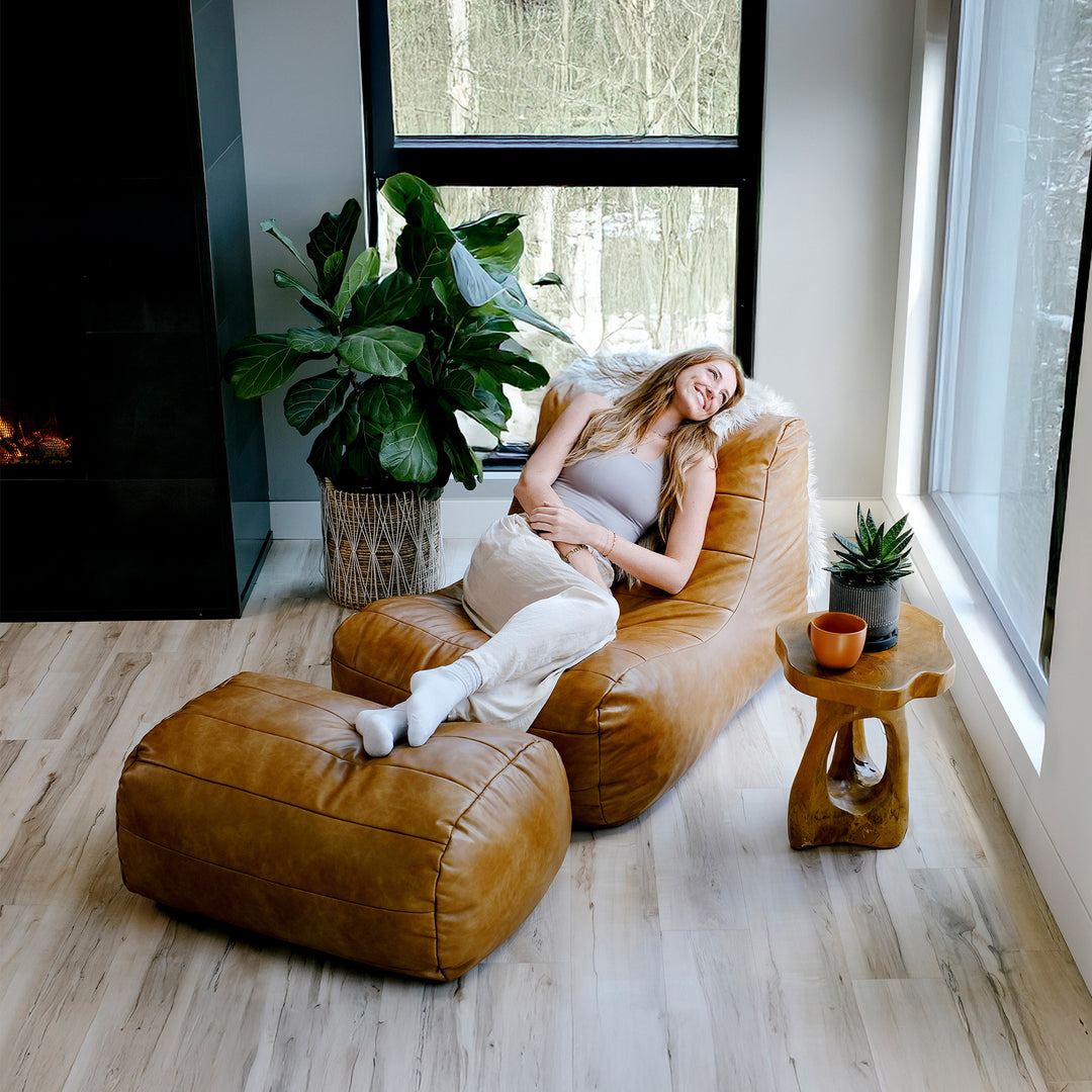 Woman sitting on a brown bean bag chair in a modern living room with large windows and plants. #color_copper-harbor-premium-vegan-leather