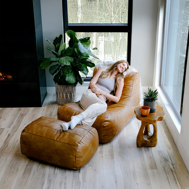 Woman sitting on a brown bean bag chair in a modern living room with large windows and plants. #color_copper-harbor-premium-vegan-leather